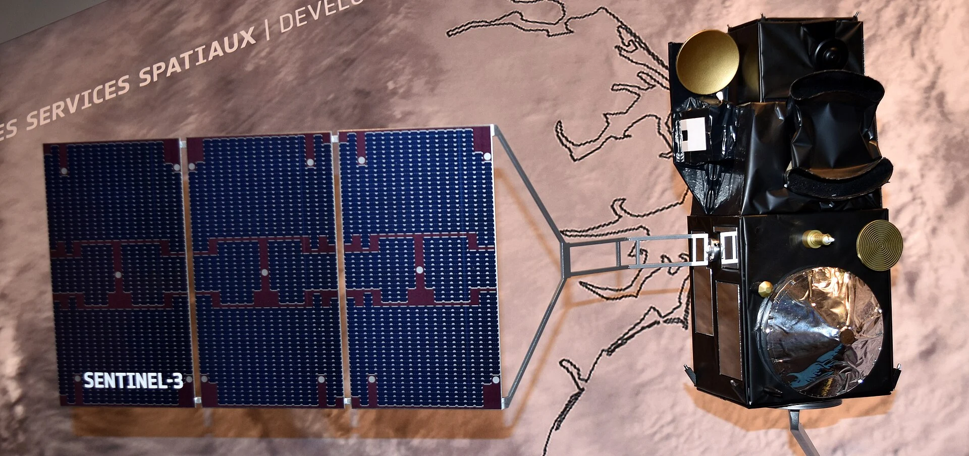 Maquette du satellite Sentinel-3 avec son panneau solaire bleu déployé devant un fond illustré